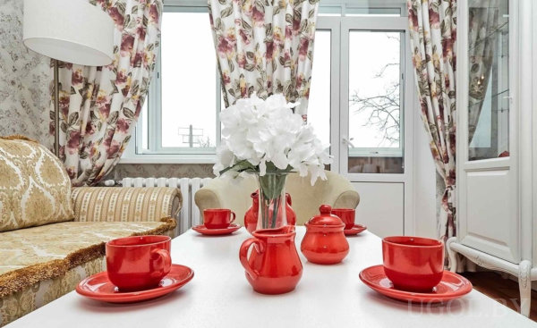 Белый стол с красным чайным сервизом, винтажный диван и цветочные шторы создают уютную атмосферу.