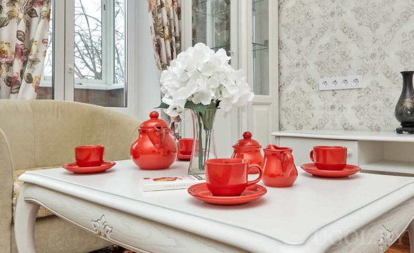 Красный чайный сервиз на белом столе, вазон с белыми гортензиями и бежевое кресло на фоне цветочных штор.