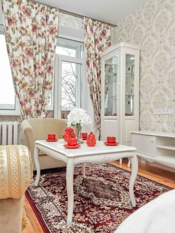 Белый резной стол на красно-бежевом ковре, красный сервиз и цветочные шторы в уютной обстановке.