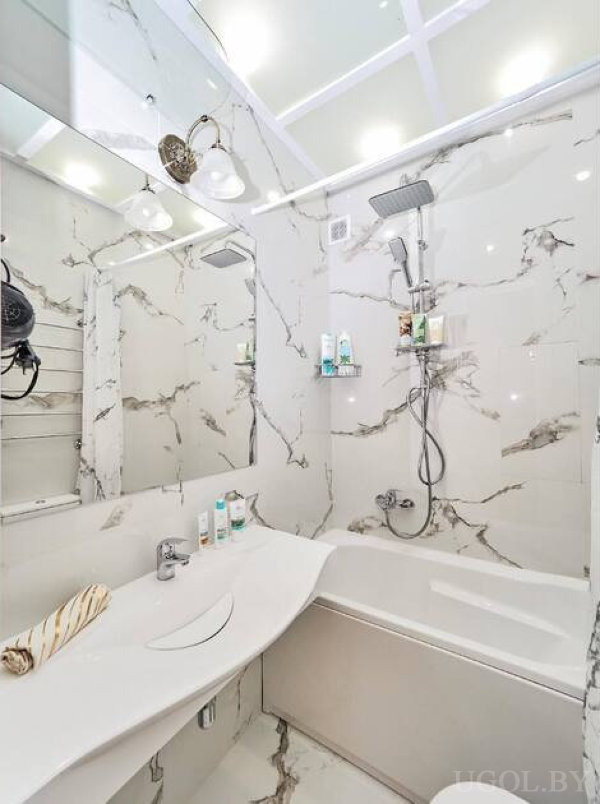 Белая ванна, зеркальная стена, подвесной душ и мраморные плитки создают стильный интерьер.