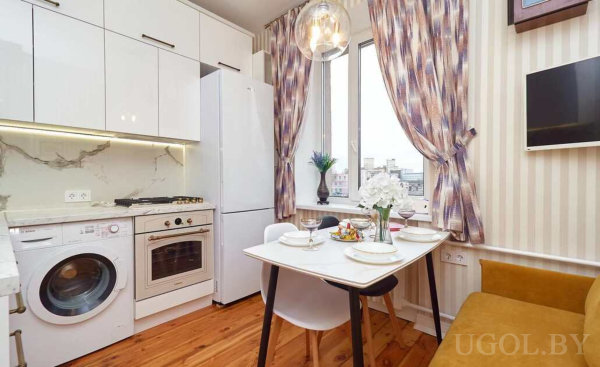 Белая кухня с техникой, стол на троих с сервировкой и горчичная скамья у окна с цветочными шторами.