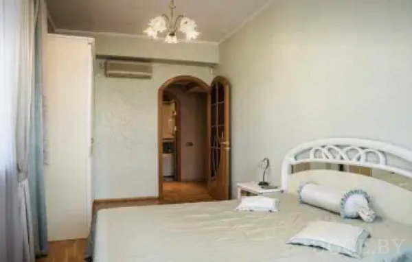 Уютная спальня с белой металлической кроватью, пастельными стенами и видом на соседнюю комнату через арочный проём. Над дверью установлен кондиционер, интерьер дополнен люстрой и деревянным полом.