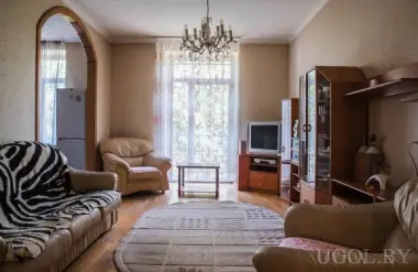 Традиционно оформленная гостиная с двумя креслами, диваном с покрывалом в зебровый принт, люстрой и большим окном с тюлем. В арке видна кухня с холодильником, на стене — ТВ и декоративные элементы.