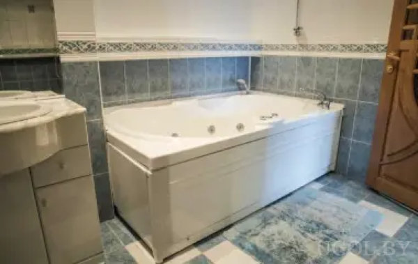 Санузел с просторной ванной с гидромассажем, синими и белыми плитками, тумбой с раковиной и деревянной дверью. Над плиткой — декоративный бордюр, интерьер выглядит чистым и обновлённым.