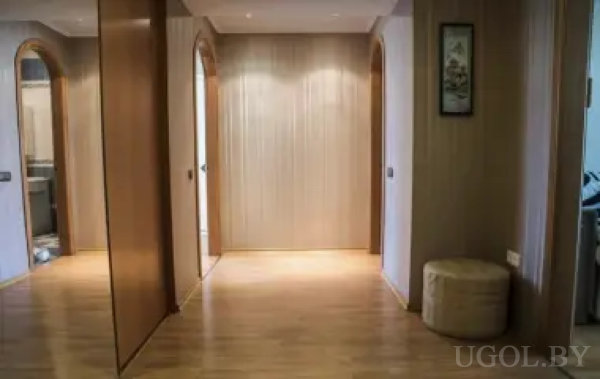 Просторный коридор с деревянным полом и стеновыми панелями, тремя арочными проходами и мягким пуфом. На стене — картина, освещение встроено в потолок, создавая мягкий свет.