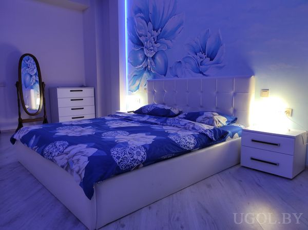 Кровать с белым изголовьем и синим покрывалом, вертикальная подсветка, панно с цветами, белая мебель и овальное зеркало.