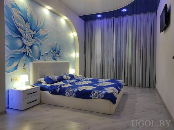 Кровать с белым изголовьем и синим покрывалом, настенные лампы, панно с цветами, глянцевый потолок и телевизор на стене.