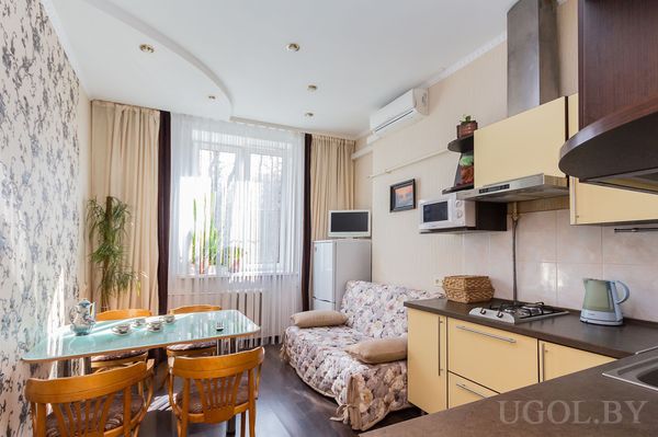 Жёлтая кухня, стеклянный стол, четыре стула, диван с цветочным узором, телевизор, холодильник и комнатные растения.