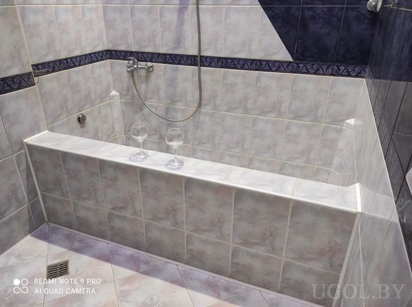 Прямоугольная ванна, два бокала на бортике, серо-мраморная плитка, душ и декоративная тёмная вставка.