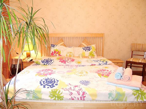 Уютная спальня с деревянной кроватью, ярким цветочным покрывалом, тремя свернутыми полотенцами и зелёным растением.