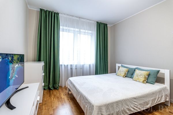 Светлая спальня с белой кроватью, зелёными и жёлтыми подушками, телевизором на комоде и шторами в тон.