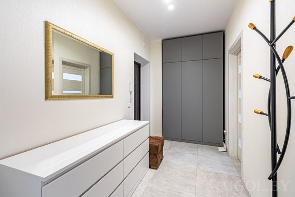 Современная прихожая с белой тумбой, зеркалом в золотой раме и встроенным серым шкафом.