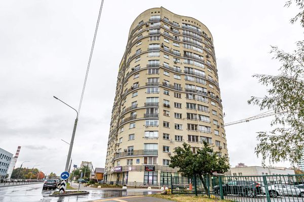 Многоэтажный дом с изогнутым фасадом, балконами, магазинами и мокрой улицей на фоне пасмурного неба.