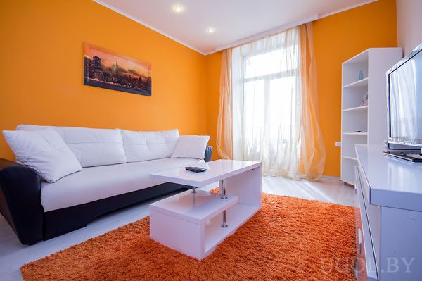 Современная комната с бело‑чёрным диваном, оранжевым ковром, журнальным столиком и телевизором на белой тумбе.