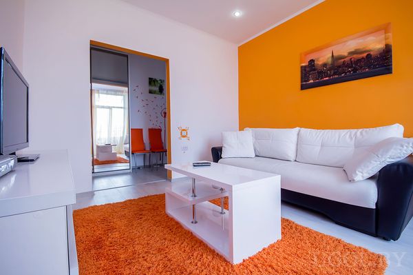 Интерьер с оранжевой стеной и диваном; через дверной проём видны оранжевые стулья и цветочные наклейки.