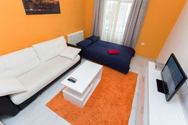 Жилая зона с оранжевой стеной, бело‑чёрным диваном, столиком и кроватью с тёмно‑синим бельём.