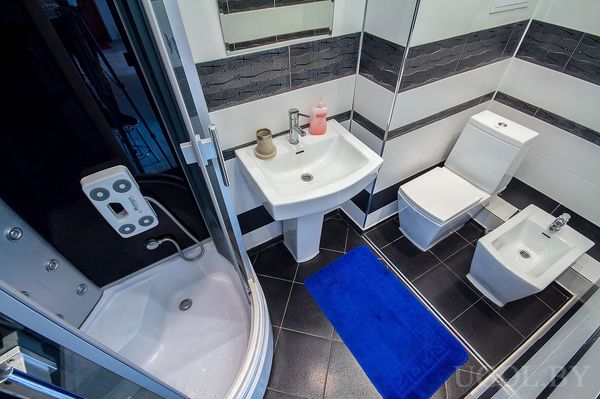 Интерьер с угловой душевой кабиной, раковиной, унитазом и биде, с чёрной плиткой пола и синим ковриком.