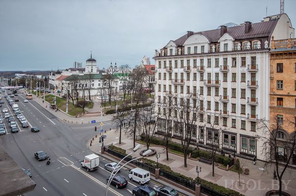 Панорама многополосной улицы с парковой зоной и архитектурой с куполами и балконами на фоне пасмурного неба.