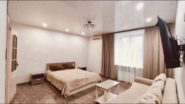 Кровать с бежевым покрывалом, диван напротив, телевизор на стене, окно с тюлем и шторами.