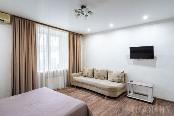 Бежевый диван с подушками, кровать на переднем плане, телевизор на стене, окно с драпировкой.