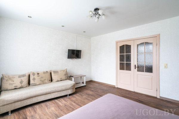 Диван с подушками, ТВ на стене, стеклянные двери с розовой рамой, тёмный ламинат и светлый потолок.