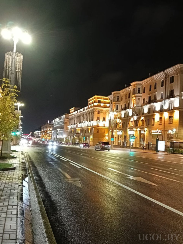 Мокрая дорога, классические здания с тёплой подсветкой, машины, деревья и фонари.