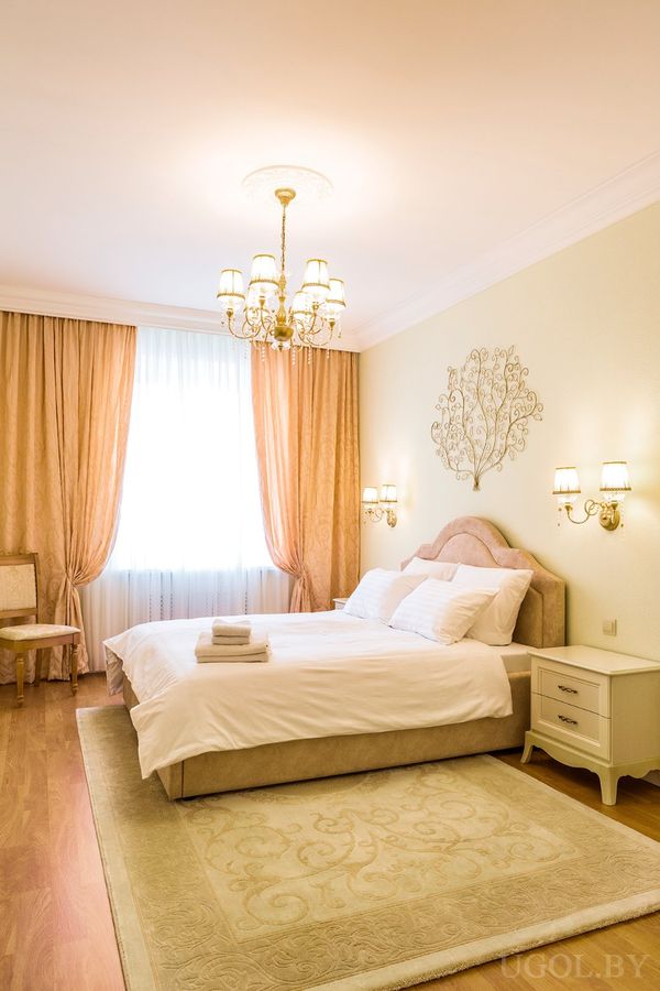 Светлая спальня с кроватью в белом белье, декоративной панелью над изголовьем, бежевыми шторами и люстрой.