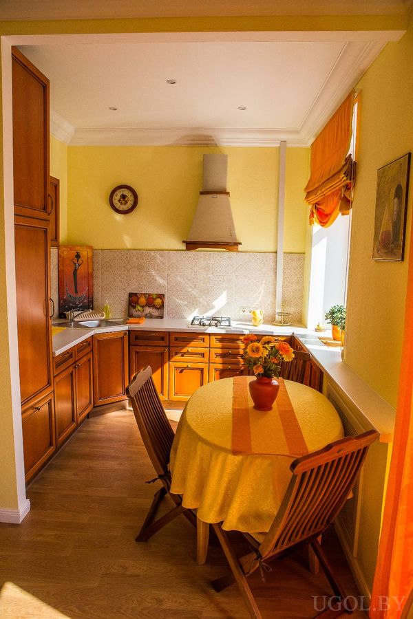 Уютная кухня с деревянными шкафами, плитой, круглым столом с жёлтой скатертью и оранжевыми шторами.