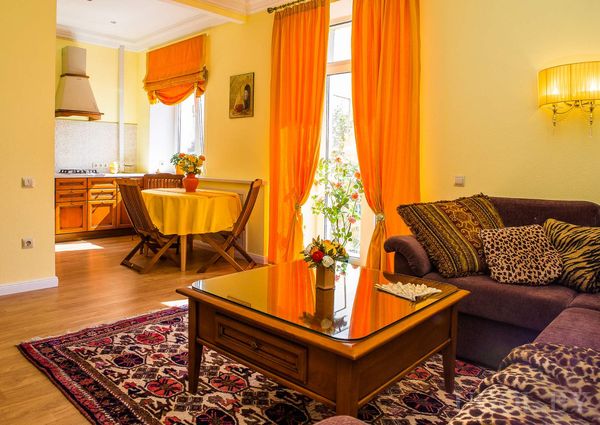 Комната с фиолетовым диваном, круглым столом с жёлтой скатертью, ковром с узором и кухней на заднем плане.