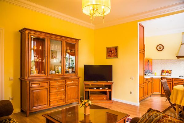 Тёплая гостиная с жёлтыми стенами, деревянным шкафом‑витриной, телевизором и круглым столом с жёлтой скатертью.