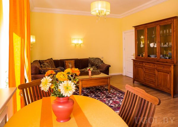 Комната с коричневым диваном, круглым столом с жёлтой скатертью, большим деревянным шкафом и ковром с узором.