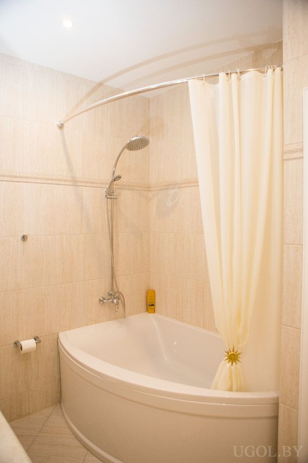 Компактная ванная с угловой ванной, бежевой шторкой, светлой плиткой и настенным душем.