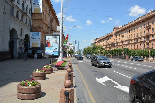 Улица с машинами, деревьями и зданием с арочными окнами и зелёными навесами на первом этаже.
