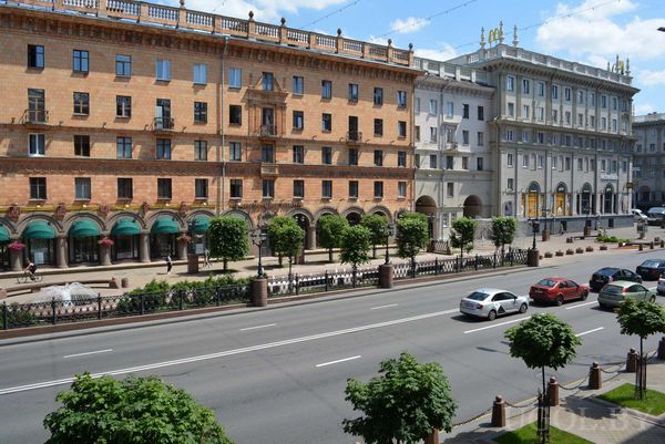 Широкая улица с машинами, пешеходными дорожками и большими зданиями в классическом стиле с колоннами и арочными окнами.