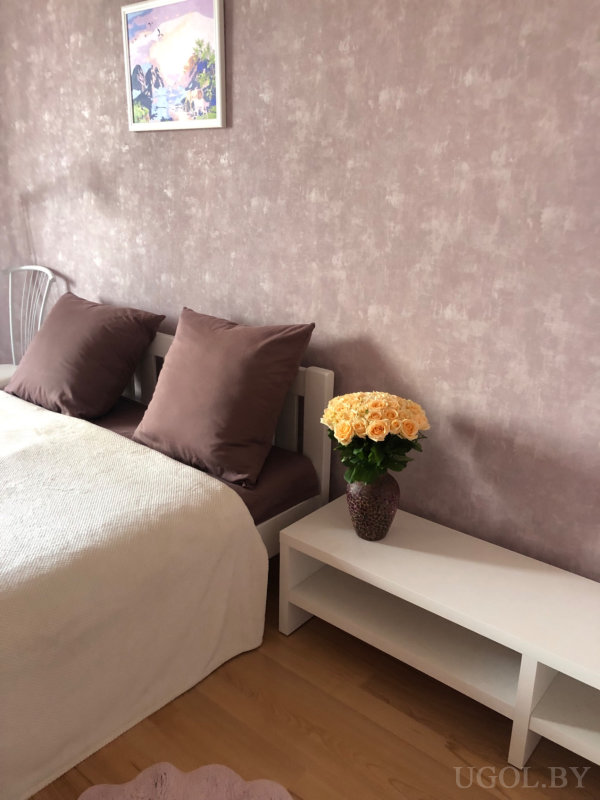 Белая кровать, светло-бежевая накидка, ваза с персиковыми розами и картина над изголовьем.