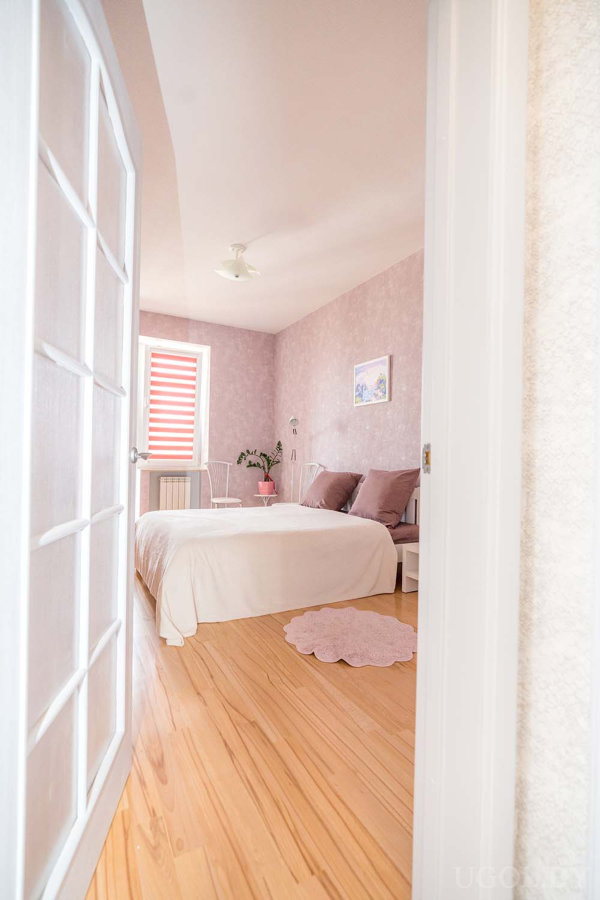 Мауфовые подушки, белое покрывало, столик с растением у окна и красно-белые жалюзи.