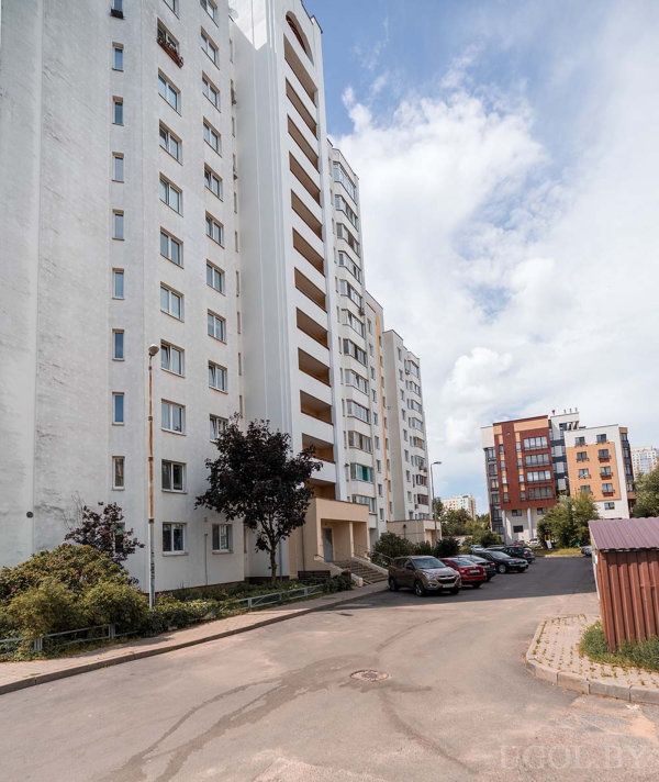 Белые и бежевые фасады, озеленение, припаркованные машины и частично облачное небо.