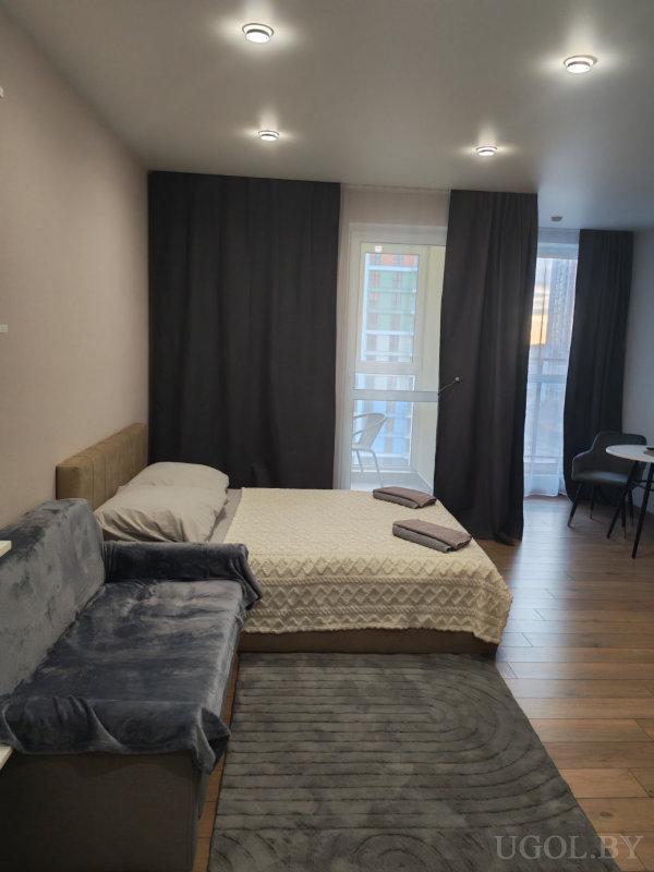 Уютная квартира-студия с кроватью, диваном, ковром и обеденной зоной у окна с выходом на балкон.