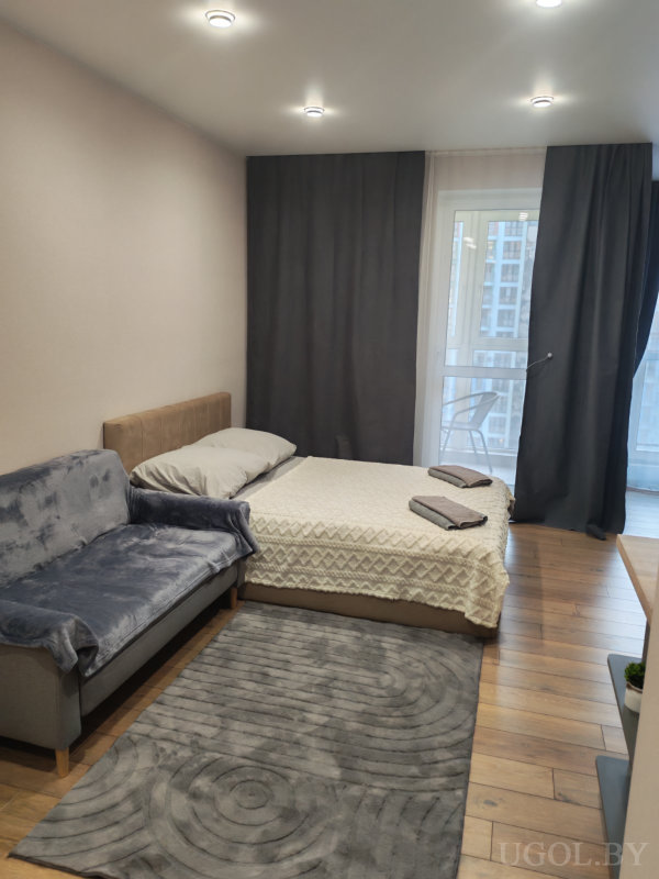 Светлая комната с кроватью, серым диваном и ковром, оформленная в спокойных тонах с видом на балкон.