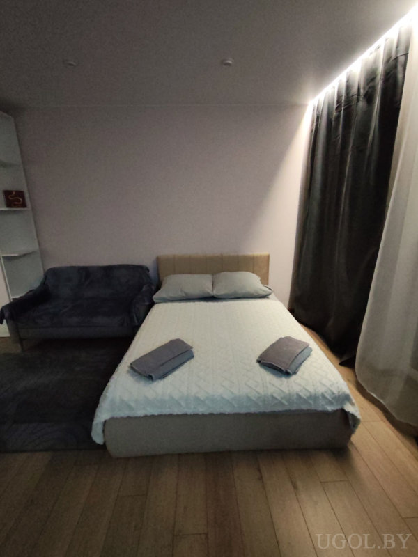 Комната с кроватью, диваном, открытым стеллажом и плотными шторами, оформленная в нейтральных тонах.