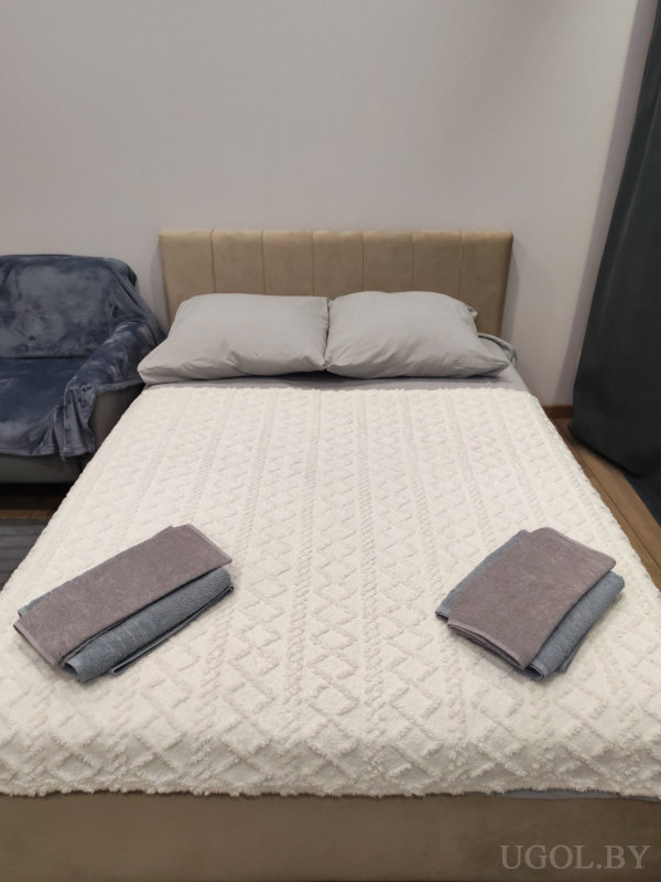 Кровать с белым покрывалом и серыми полотенцами, рядом кресло с пледом и плотные шторы.