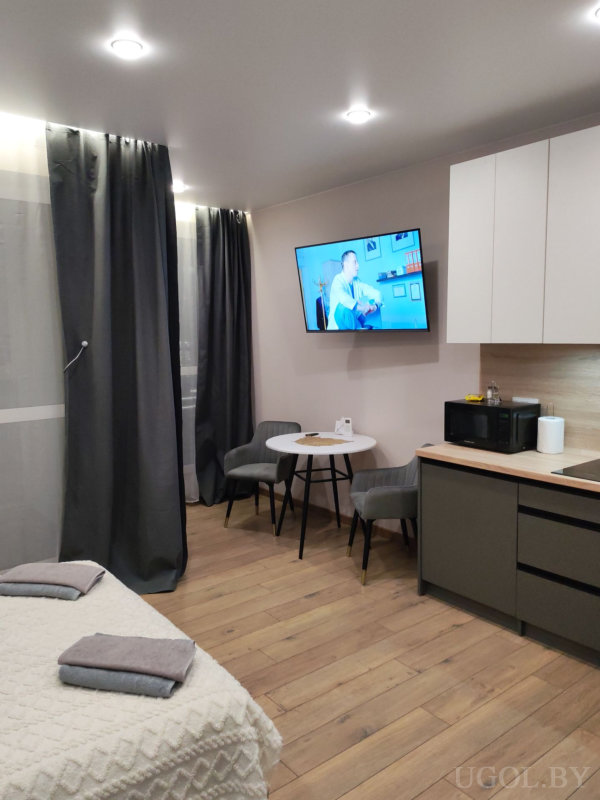 Кровать, обеденный стол, телевизор и кухня с белыми и тёмными фасадами, оформленные в современном стиле.