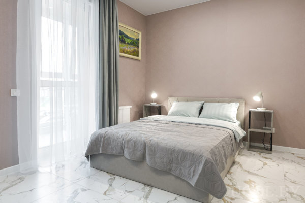 Кровать с серым стёганым покрывалом и голубыми подушками, две тумбочки с лампами, мраморный пол и окно с тюлем и серыми шторами.