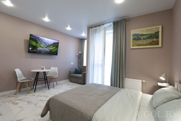 Кровать с бело‑серым текстилем, небольшой круглый стол с двумя стульями, телевизор на стене, кресло у окна и мраморный пол.