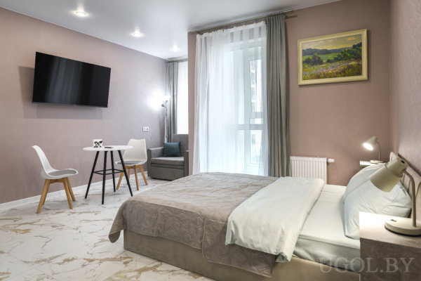 Кровать с бело‑серым текстилем, круглый стол с двумя стульями, телевизор на стене, серое кресло у окна и мраморный пол.