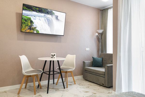 Круглый белый стол с двумя стульями, телевизор на стене с изображением водопада, серое кресло с бирюзовой подушкой и торшер у окна.