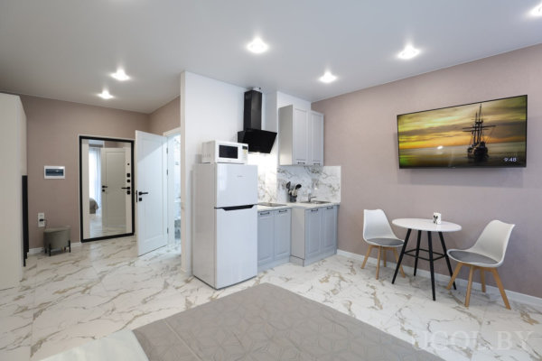 Компактная кухня со светло‑серыми фасадами, круглым столом с двумя белыми стульями и телевизором на стене с изображением корабля на закате.