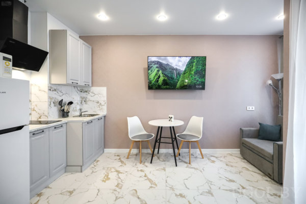 Светлая кухня с мраморным фартуком, круглым столом и двумя белыми стульями, рядом серый диван и телевизор с пейзажем гор.