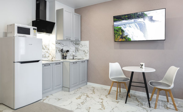 Кухня со светло‑серыми шкафами, белым холодильником и круглым столом с двумя стульями, на стене телевизор с изображением водопада.