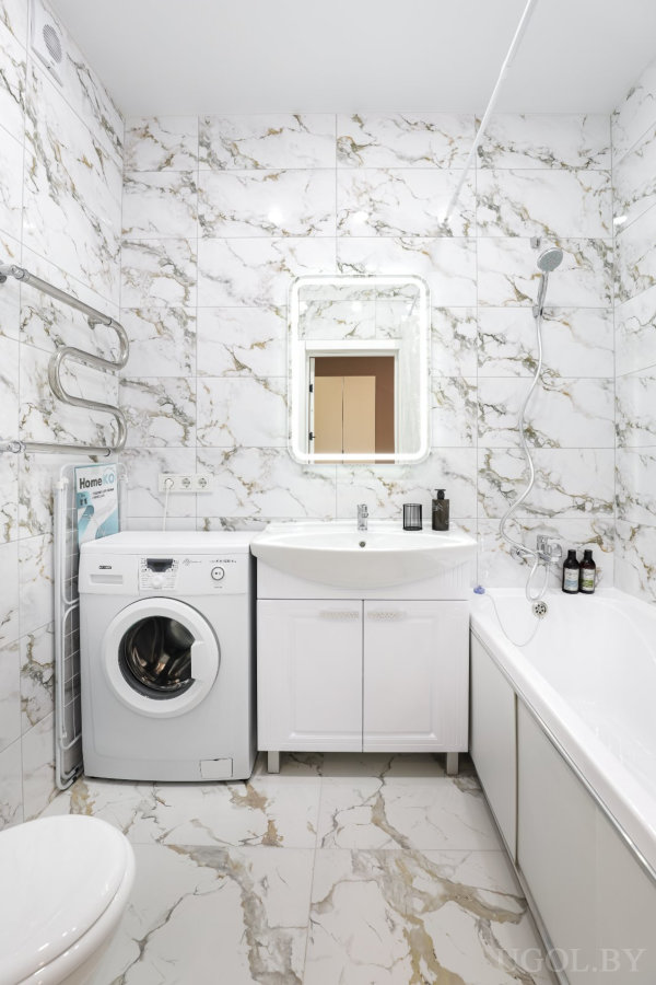 Современная ванная с мраморными стенами и полом, стиральной машиной, раковиной с тумбой, подсвеченным зеркалом и ванной с душем.